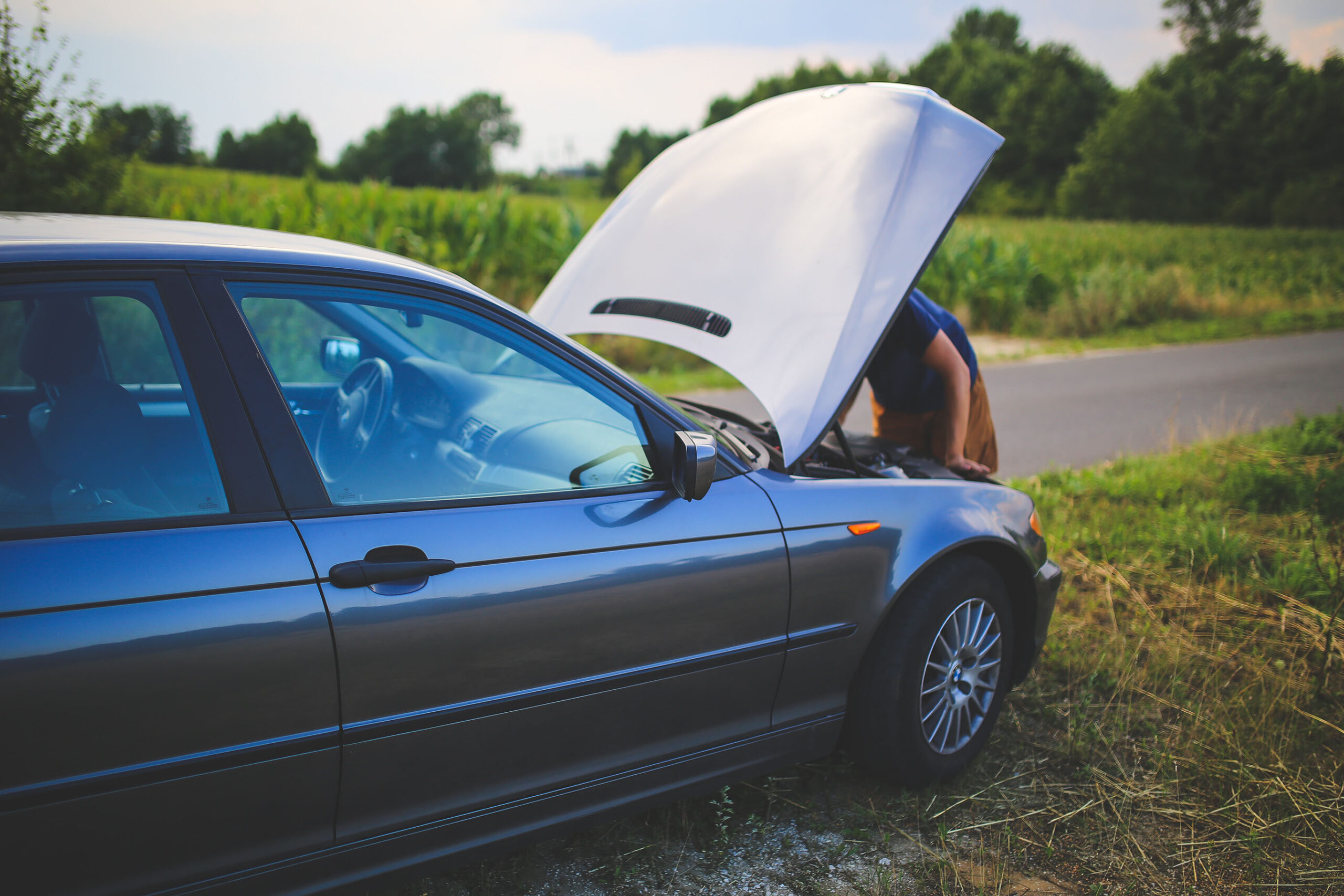 In einer ländlichen Gegend steht ein Auto am Straßenrand mit offener Motorhaube während sich eine Person über den Motor beugt.