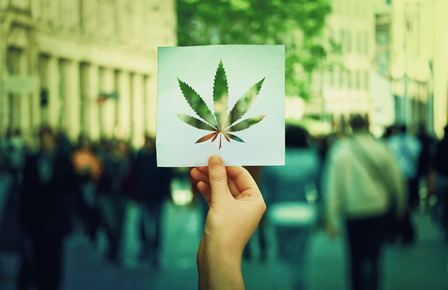 Im Vordergrund hält eine Hand eine Papier mit einer ausgeschnittenen Silhouette eines Cannabis-Blatts hoch. Im Hintergrund laufen Menschen auf der Straße.