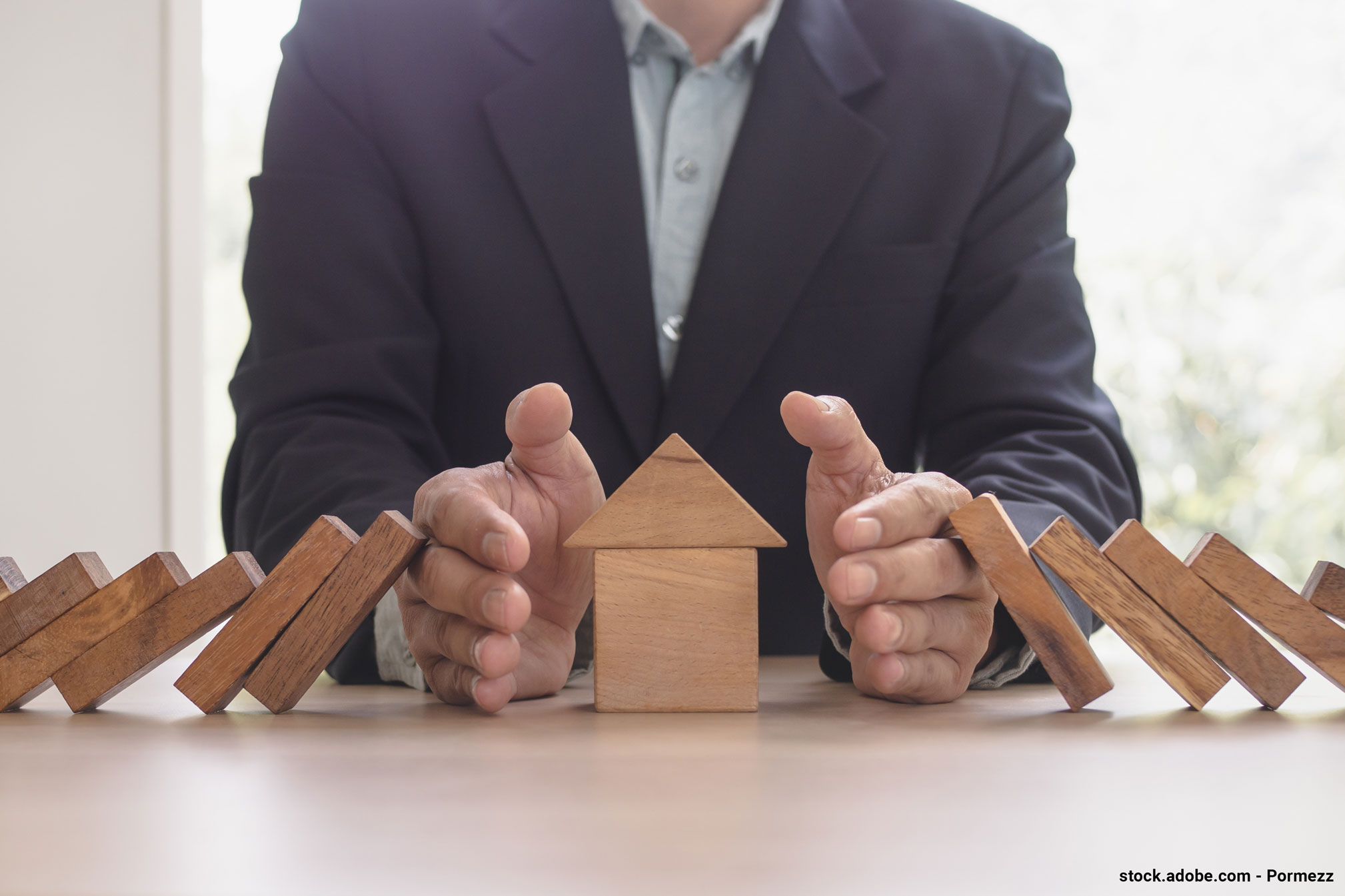 Zwei Hände schützen ein kleines Haus aus Bauklötzen, während andere Bauklötze drohen, auf das Haus zu fallen.